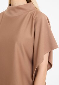 Brown, high-neck top featuring short sleeves, a relaxed fit, and a smooth texture. Material appears soft and lightweight.
