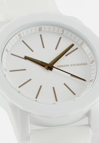 Reloj de silicona blanca con una esfera redonda, diseño minimalista, marcadores de hora dorados, dos agujas doradas y un logo de 'Armani Exchange'.
