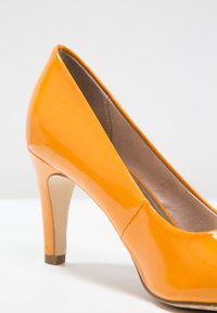 Chaussure à talon haut orange brillant, inclinée pour montrer le côté et le talon arrière sur une surface blanche avec un fond gris.
