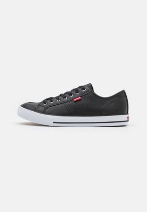 Sneakers in pelle nera con suola in gomma bianca, punta rotonda e strisce nere a contrasto. Presentano occhielli metallici e una piccola etichetta con logo rosso.