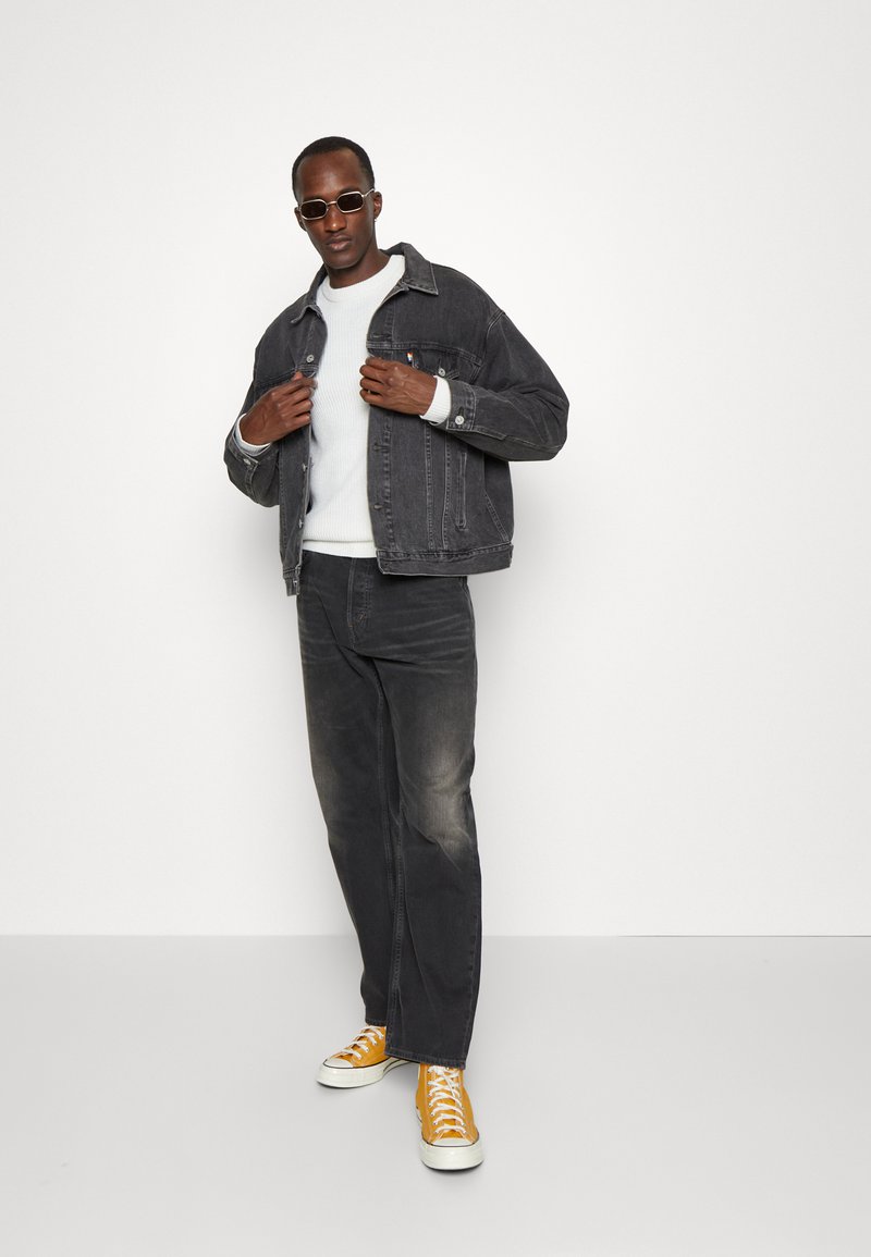 Hombre con chaqueta de mezclilla oscura, suéter blanco, jeans oscuros, zapatillas amarillas de caña alta y gafas de sol, de pie contra un fondo blanco liso.