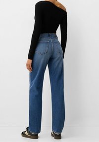 Černý top s odhalenými rameny a dlouhými rukávy, zkombinovaný s modrými džíny s vysokým pasem, které mají rovný střih a viditelné zadní kapsy.