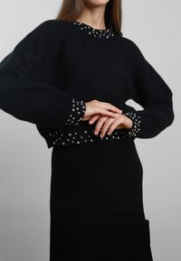 Maglione nero lavorato a maglia con una texture a coste, dotato di polsini e orlo decorati con piccole borchie argentate. Progettato con una vestibilità rilassata.