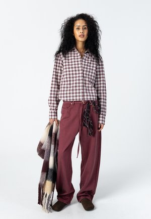 Vrouw met krullend haar, gekleed in een geruit blouse met knopen en een losse bordeauxrode broek, die een grote geruite sjaal met franjes vasthoudt.