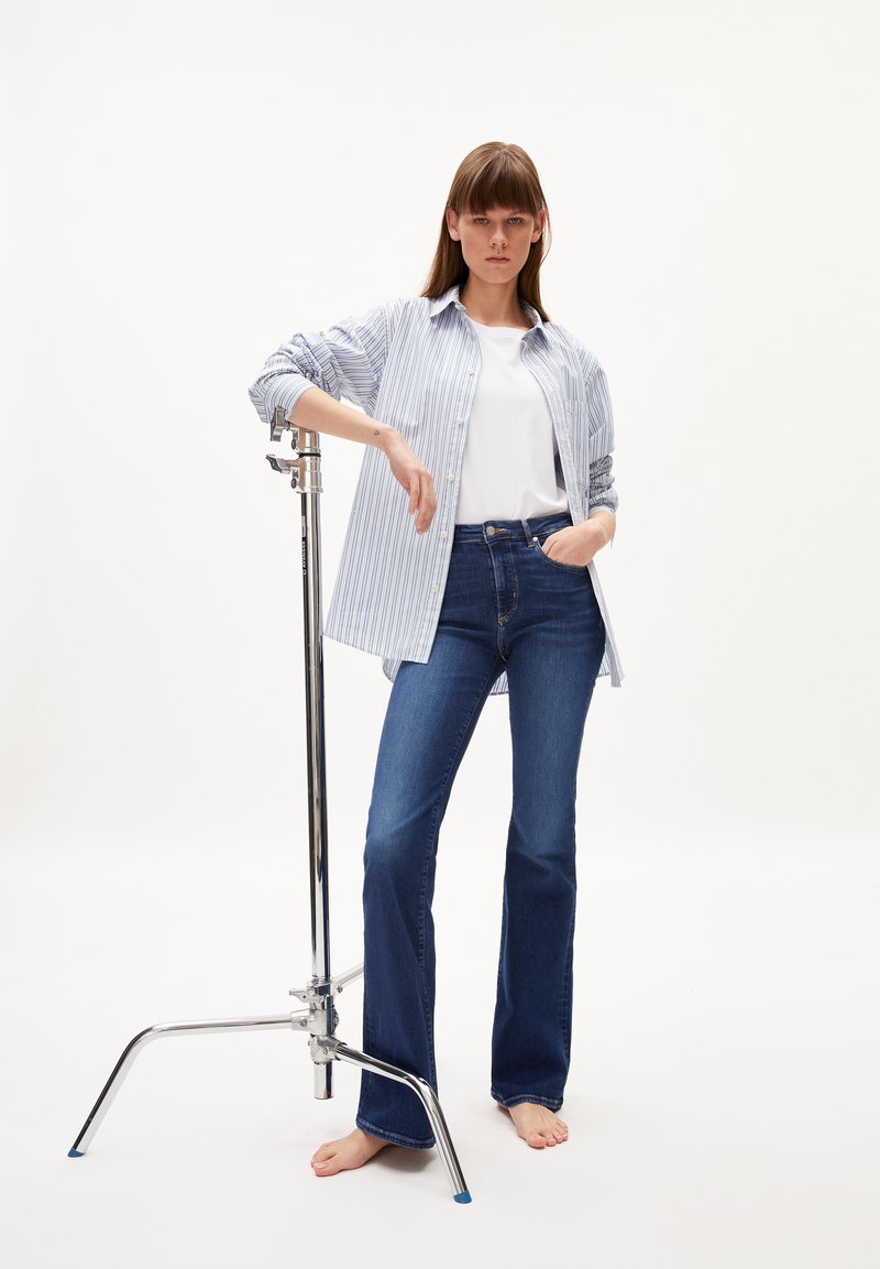 Gestreiftes blau-weißes Hemd, das über einem weißen T-Shirt getragen wird, kombiniert mit dunkelblauen Schlagjeans. Das Model steht barfuß neben einem chromfarbenen Kleidungsständer.