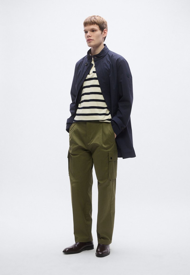 Navy lightweight jacket over a striped beige and navy shirt, paired with olive cargo pants, and brown leather shoes.