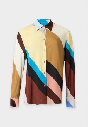 Kvinde med glat hår iført en flerfarvet langærmet trøje og en lang vinrød nederdel med beige spidse hæle, stående mod en ensfarvet baggrund.
