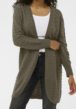 Kvinde iført en olivengrøn kabelstrikket cardigan over en hvid top og sorte jeans, justerer ærmerne på cardiganen med hænderne.