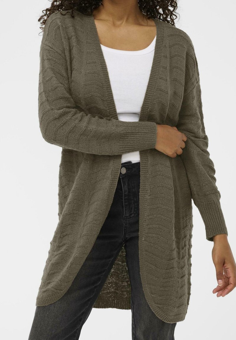 Femme portant un cardigan vert olive tricoté en torsades par-dessus un haut blanc et un jean noir, mains ajustant les manches du cardigan.