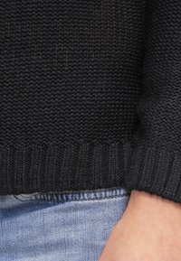 Černý pletený svetr s žebrovaným lemem a texturovanou látkou, částečně viditelný nad modrými džínami, viditelná pouze ruka.