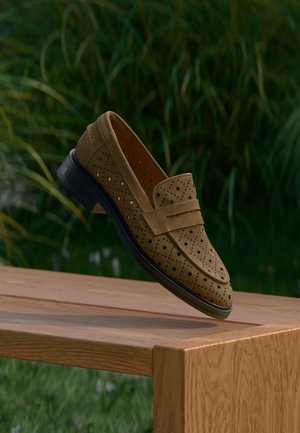 Braune Wildleder-Loafers mit perforiertem Muster und dekorativem Riemen, die an einer Holzoberfläche lehnen, vor unscharfem grünem Hintergrund.