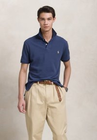 Noor mees, seljas tumesinine polo särk, millel on tõstetud krae, beežid plissee püksid, punutud pruun vöö ja käekell, seisab käsi taskus.