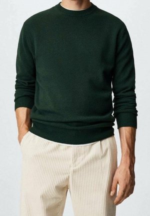 Homme portant un pull en maille vert foncé avec des poignets côtelés et un pantalon côtelé de couleur crème, les mains détendues le long du corps.