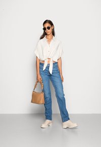 Blusa branca de mangas curtas com nó; jeans de perna reta azul claro; sapatos de plataforma creme; mala castanha; óculos de sol pretos; texturas suaves.