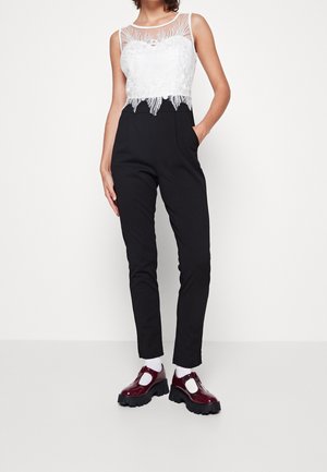 Schwarzer Jumpsuit mit einem weißen Spitzenoberteil mit Blumendesign, ärmellosem Design und gezacktem Saum; kombiniert mit bordeauxroten Lackschuhen.