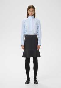 Blouse bleu clair à manches longues avec col montant, associée à une jupe grise évasée. Des collants noirs et des chaussures plates complètent le look.