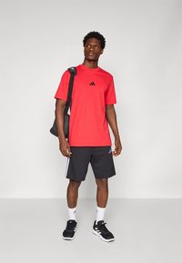 Raudona sportinė marškinėliai su juodu logotipu, derinami su juodais šortais su baltais dryžiais. Modelis dėvi juodus batus ir nešioja tamsią pečių krepšį.