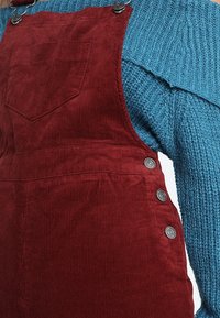 Röd kordflorseoverallkjol med framficka och metallknappar på sidan, bärs över en blå stickad tröja.