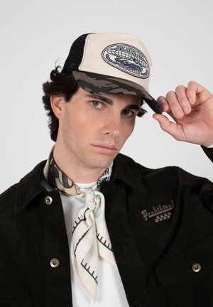 Jonge man met een beige en camouflagepet met de tekst "Prohibited Racing Club", een zwarte jas met knopen en een patroon sjaal om de nek, kijkt naar de camera.