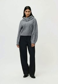 Femme se tenant debout, portant un pull en tricot gris à col haut et un pantalon noir à jambes larges, devant un fond clair.