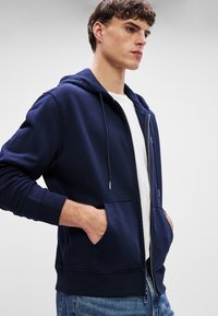 Hanorac cu fermoar de culoare bleumarin, dintr-un material moale, având un buzunar frontal, glugă cu șnur și manșete ribbate. Purtați peste un tricou alb.