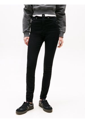 Personne portant un jean skinny noir, des baskets noires à semelles claires et un sweat gris court, debout devant un fond blanc uni.