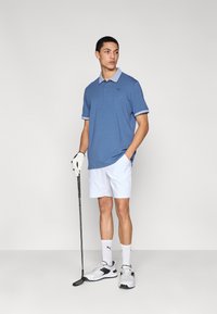 Μπλε πόλο μπλούζα με ανοιχτό μπλε λεπτομέρειες, λευκό σορτς, γάντι γκολφ και παπούτσια. Το μοντέλο στέκεται με ένα γκολφ ρόπαλο, παρουσιάζοντας μια αθλητική εμφάνιση.