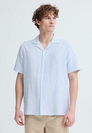 Jonge man met krullend haar, gekleed in een lichtblauw-wit gestreept overhemd met korte mouwen en beige broek, kijkt naar rechts.