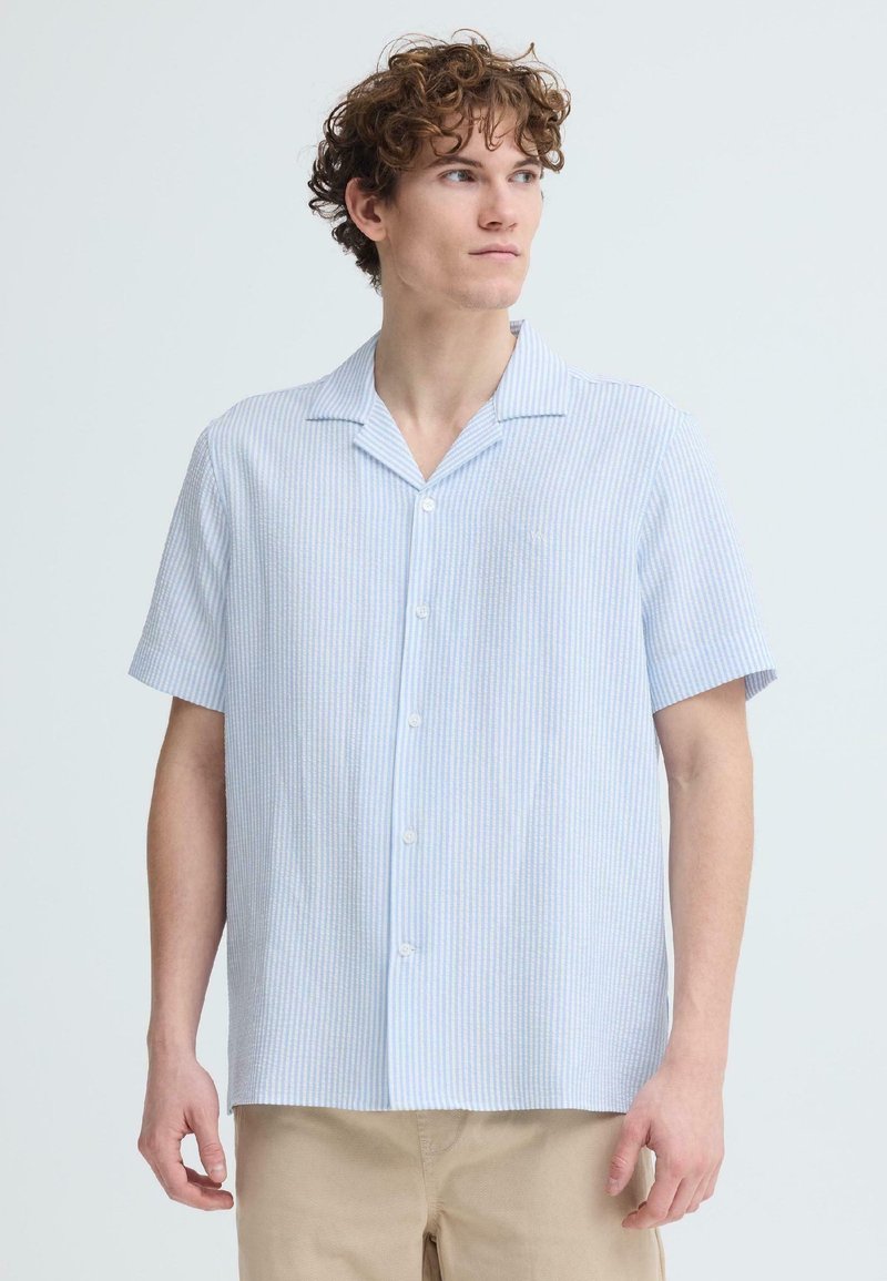 Mladý muž s kučeravými vlasmi, oblečený v svetlomodrej a bielej pruhovanej košeli s krátkym rukávom a béžových nohaviciach, pozerajúci sa doprava.