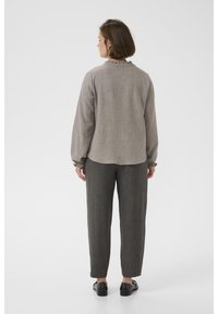 Femme debout, vue de dos, portant un chemisier à manches longues gris clair avec des poignets à volants et un pantalon droit gris foncé avec des chaussures noires.