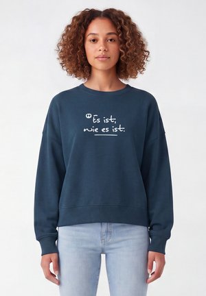 Vrouw met krullend haar die een marineblauwe trui draagt met de tekst "ts ist, wie es ist" in het wit en lichtblauwe spijkerbroek, staand tegen een egale achtergrond.
