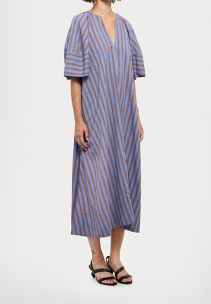 Kobieta w luźnej sukience do kolan z niebiesko-brązowymi ukośnymi paskami, z krótkimi rękawami, w czarnych sandałach z odkrytymi palcami.