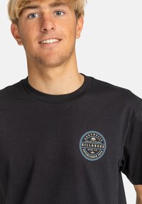 Black cotton t-shirt featuring a round logo patch on the left chest with blue and gold accents, short sleeves, and a crew neckline.