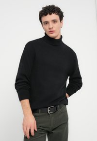 Pull à col roulé noir en tissu maille doux, avec manches longues et poignets côtelés, assorti à un pantalon en velours côtelé vert olive.