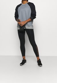 Grå och svart sweatshirt med raglanärmar och rundad nederkant; tillsammans med svarta leggings med utskurna detaljer längs sidorna. Svarta sneakers.
