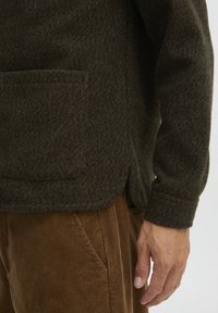 Suéter de lana verde oscuro con un bolsillo lateral, tejido texturizado y dobladillo redondeado. Combinado con pantalones de pana marrones, que muestran líneas verticales.
