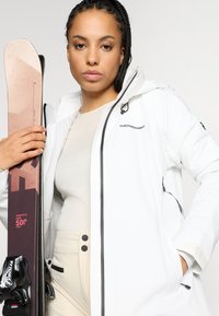 Casaco impermeável branco com detalhes e fecho preto, combinado com calças bejes. Uma ski com um design rosa e preto está sendo segurada.