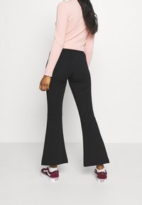 Woman wearing black flared pants, light pink long-sleeve top, and maroon and white sneakers, standing against a plain white background.