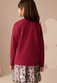 Maglia lunga color bordeaux abbinata a una gonna con paillettes che presenta motivi ondulati in rosa, blu e oro. Tessuto morbido e vestibilità comoda.