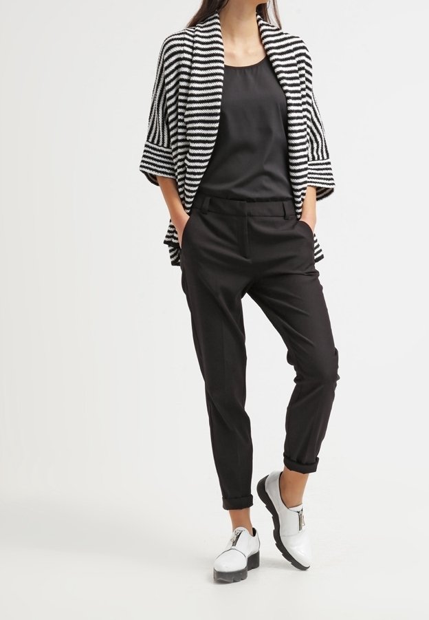 Femme portant un pantalon noir et un haut sans manches, un châle rayé noir et blanc, et des chaussures compensées blanches, debout les mains dans les poches.