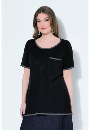 Frau mit langen braunen Haaren trägt ein schwarzes kurzärmeliges Oberteil mit silbernem Besatz und Tasche, kombiniert mit einem dunklen Jeansrock, steht vor einer weißen Wand.