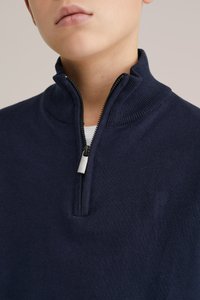 Tumši zils džemperis ar rāvējslēdzēju un fakturētu apkakli, aprīkots ar ceturtdaļas garuma rāvējslēdzēju un nelielu metāla rāvējslēdzēja cilni.