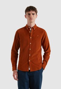 Camicia in corduroy arancione bruciato con colletto button-down, bottoni bianchi e un piccolo logo ricamato sul petto. Vestibilità casual, maniche lunghe.