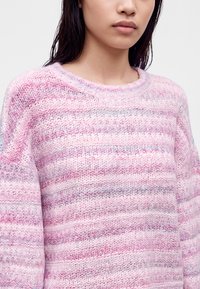 Jeune personne portant un pull tricoté à rayures roses et lavande, visible des épaules au menton sur un fond uni.