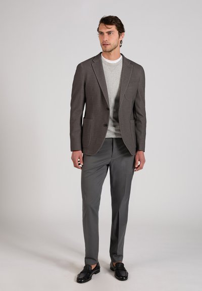 Homme portant un blazer gris foncé, un pull gris clair, un pantalon gris assorti et des mocassins noirs, debout devant un fond uni.