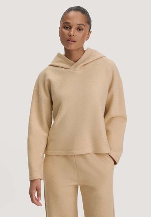 Jonge vrouw met naar achter getrokken haar, gekleed in een bijpassende beige hoodie en broek, staand met één hand in de zak tegen een lichte achtergrond.