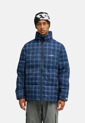 Jonge man draagt een blauw geruite donsjas met rits dicht en een zwarte gebreide muts met wit logo op een lichte achtergrond.