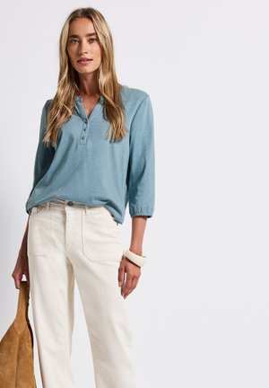 Haut bleu clair à manches longues avec un col à boutons, associé à un pantalon crème taille haute et un sac beige. Présente une texture lisse.