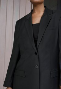 Černý strukturovaný blazer s jedním knoflíkovým zapínáním, skloněnými klopami a dvěma předními kapsami, vyrobený z hladkého materiálu.