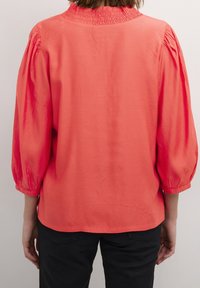 Donna che indossa una blusa corallo con maniche a tre quarti arricciate e colletto arricciato, abbinata a pantaloni neri, in piedi su uno sfondo neutro.